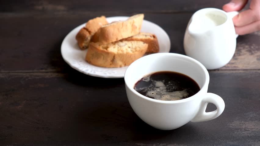 Adding milk cream into black coffee. Coffee with milk and italian cookies Cantucci. Wooden table background | Shutterstock HD Video #1106384911