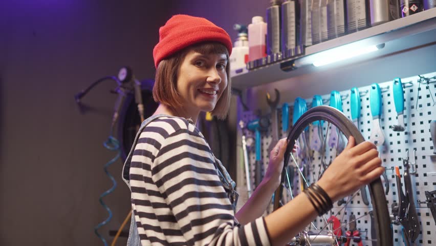 Female master repairing bicycle wheel in workshop. Cycling mechanic looking at camera and smiling. Woman fixing bicycle spokes at workshop. Preety female craftworker near rack with tools.