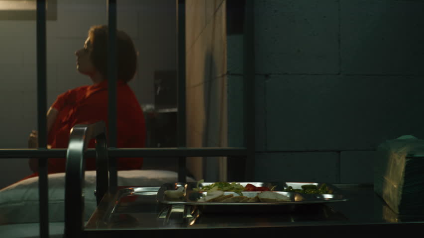 Prison worker gives food from serving trolley to female prisoner in orange uniform in jail cell. Woman criminal sits on bed, eats dinner. Imprisonment term in prison or detention center. Dolly shot. Royalty-Free Stock Footage #1106388635