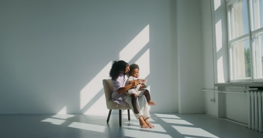 Mom with a child of African-American appearance, dressed in white clothes, playing pyramid while sitting in a chair in a white room near the window