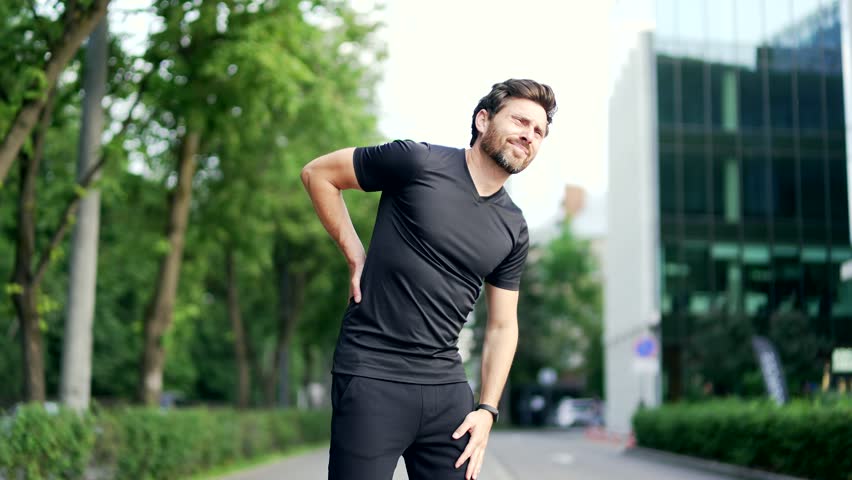 Mature adult runner suffering from back pain, sports injury, muscle spasm in urban city park. Handsome man in sportswear having back flank ache and problem after exercise jogging outside outdoors Royalty-Free Stock Footage #1106396009