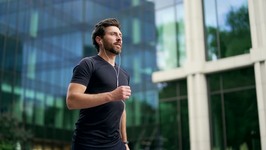 Handsome bearded male runner running on a modern urban background of buildings a city street. Sporty man jogger in headphones jogging outdoors look at result a fitness tracker bracelet or smartwatch Royalty-Free Stock Footage #1106396025