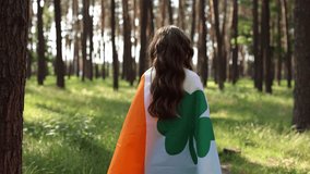 Little girl celebrating St Patricks Day. Video camera following a cute white kid covered with large Irish flag walking through a forest at sunset