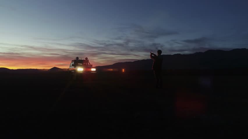 Handsome hipster man with beard stands next to SUV car and makes photos on smartphone of beautiful and inspiring mesmerizing sunset skies with purple and pink color palette