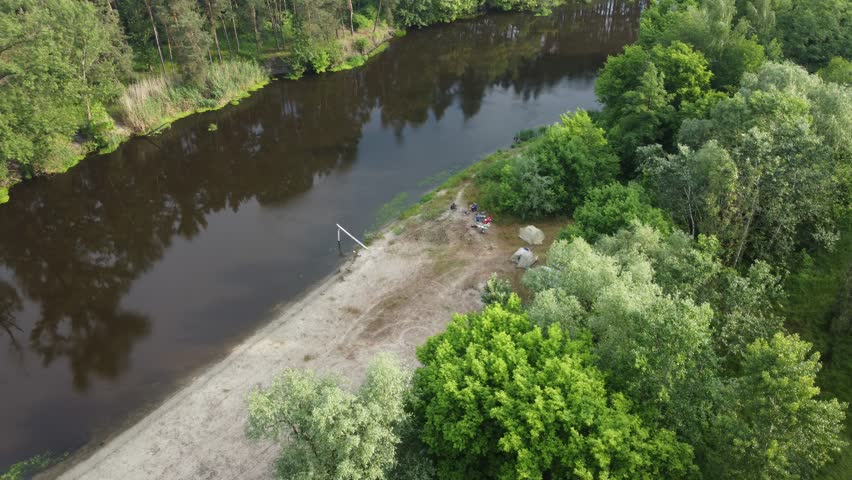 View of a small river among the forest from a drone in summer. A small group of people is visible on the shore.