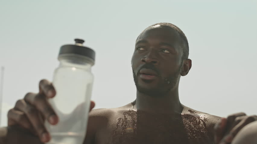 Low angle shot of muscular shirtless black man sitting outdoors on sunny day, drinking and pouring water from bottle over his head while resting after intensive physical training Royalty-Free Stock Footage #1106417201