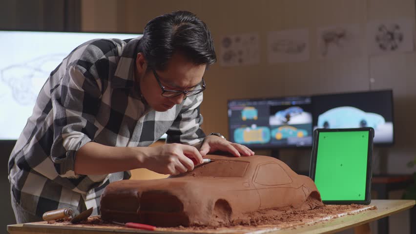 Asian Man Automotive Designer With Green Screen Tablet Using Rake Or Wire To Smooth Out The Surface And Create Details In The Sculpture Of Car Clay In The Studio
