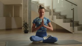 Woman is doing namaste gesture after meditation and rolling up her yoga mat. Girl in blue leggings smiling after yoga session at home
