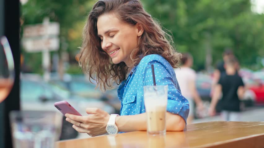A young Caucasian cheerful woman using a phone on the outdoor terrace of a cafe on a summer day.