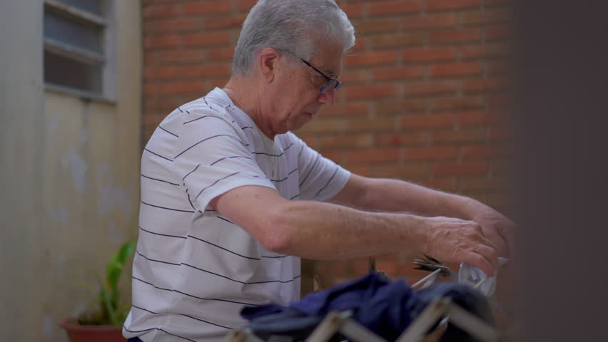 Casual candid scene of an elderly senior doing domestic household chores at home, putting clothes to dry on hanger in home backyard