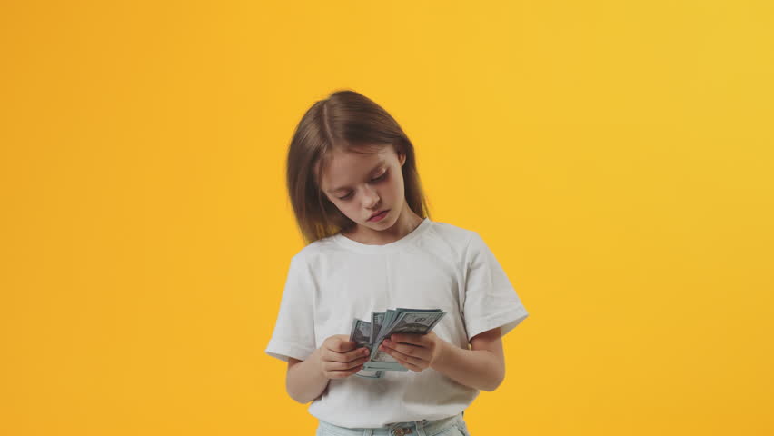 Serious Caucasian girl kid wearing white t-shirt hold fan of cash money in dollar banknotes and counting it on plain yellow background in studio