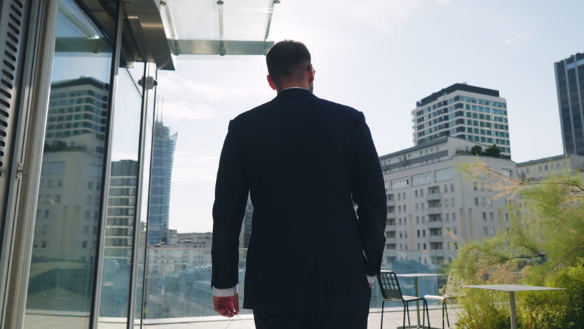 Rear back view following handsome male businessman in suit walks roof top terrace in office building overlooking modern city downtown cityscape with high-rise skyscrapers backlit with bright sunlight Royalty-Free Stock Footage #1106431609