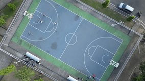 4K video footage aerial bird-eye view of a basketball court with trees. Young people are practicing Basketball game training with coach on the sport field in a blue green park area in the sunset light