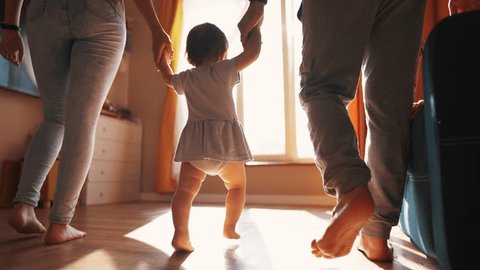 parents teach baby to walk. baby toddler taking first steps. happy family kid dream concept. parents walk with their daughter toddler indoors hold hands teach to lifestyle walk first steps วิดีโอสต็อก
