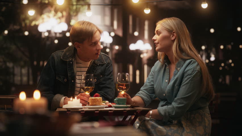 Young Man and Woman Having an Evening Date on a Terrace. Handsome Man Telling Interesting Funny Stories, Beautiful Female Enthusiastically Listening. Couple Toasting and Clinking Glasses with Wine