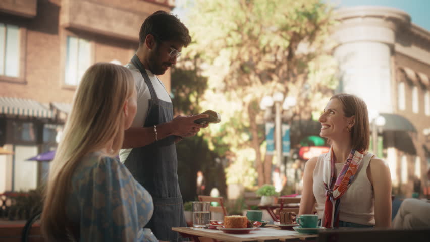 Two Female Friends Ordering Delicious Cake Desserts From a Member of the Staff in a Cafe. Handsome Multiethnic Waiter Holding a Terminal, Female Using Her Smartphone for a Contactless NFC Payment Royalty-Free Stock Footage #1106449025