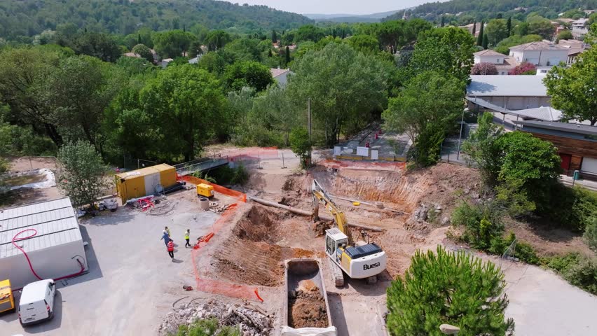 aerial drone view of a building site surrounded by trees, with a backhoe picking up the soil to put it on a truck, workers on the site, safety barriers Royalty-Free Stock Footage #1106454053