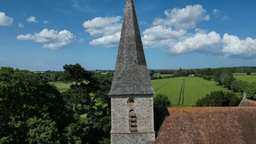 Rising push-in shot of the spire of St John the Evangelist church in Ickham, with large and sparse clouds in the blue sky.