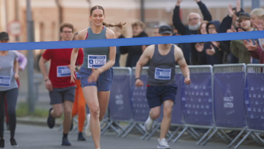 Slow Motion Portrait of Athletic Female Jogger Crossing the Finish Line in Marathon Race with the Audience Cheering. Happy Successful Woman Celebrating Winning, Feeling Empowered with her Achievement