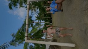 Vertical video. Aerial dynamic shot tourist happy love couple young man and girl swimsuits swing resting paradise tropical island hill covered with palm trees ocean in Asia, receding FPV sport drone