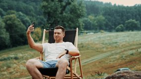 Smiling Caucasian man sits in the nature. Man holds his phone high talking with someone by video chat.