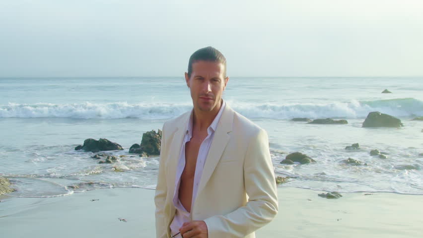 Handsome man in white suit walking by Pacific Ocean. Man puts up stylish sunglasses on the beach Royalty-Free Stock Footage #1106462907