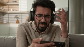 Disappointed Arabian Indian man guy watching sport game streaming on mobile phone upset by lose football match on couch in kitchen frustrated male in headphones looking at cellphone bad result loss - Powered by Shutterstock - Get 15% off with code: PIKWIZARD15