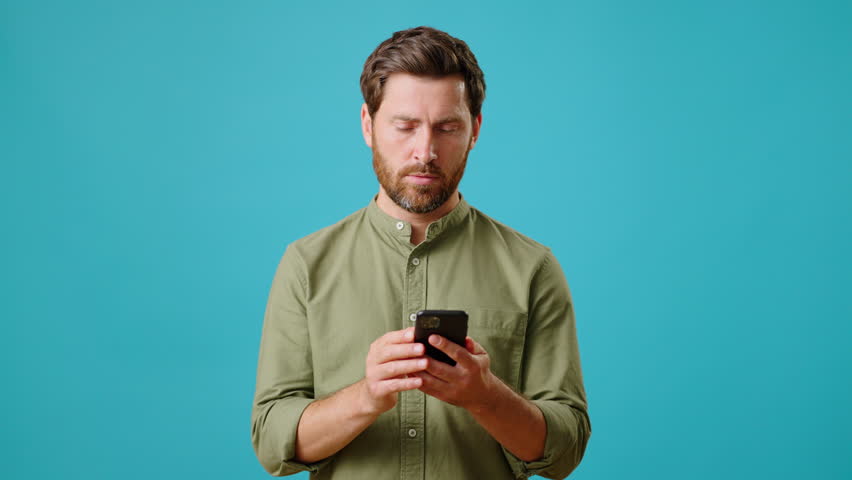 Young bearded man in green shirt smiles and points at mobile phone screen. Cheerful guy user recommends application or site on turquoise background Royalty-Free Stock Footage #1106467789