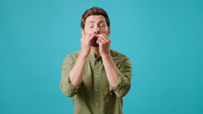 Young bearded man makes chef kisses and says Belisimo. Joyful male person praises preparation of dish smiling with satisfaction on turquoise background