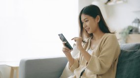 Happy young asian woman relax on comfortable couch at home, typing chat message on smartphone, smiling girl use cell phone chatting, searching information on browser wireless internet, online shopping