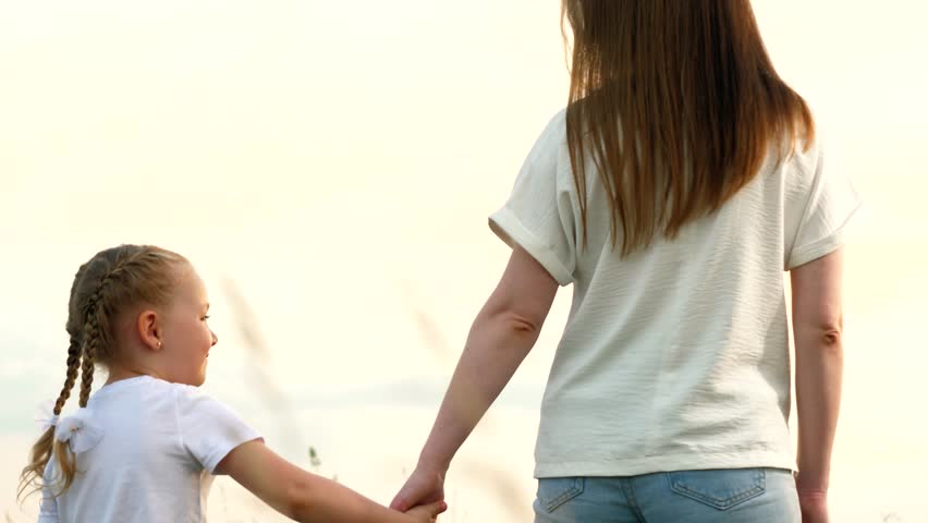 Mother and little daughter walk joining hands across field at sunset light backside view. Happy daughter holds mother hand walking together on country meadow. Mother and daughter enjoy family time