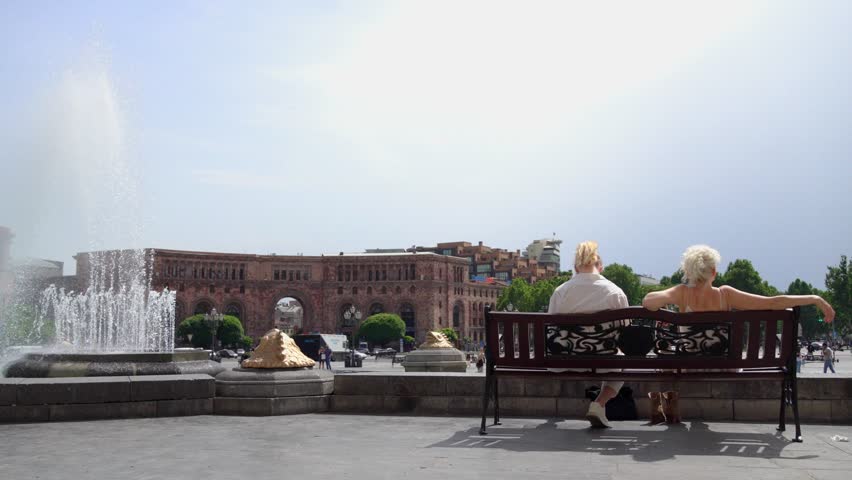 Two women sitting on bench and enjoying pleasant sunny day in city center on Republic Square in Yerevan, Armenia with fountains in backdrop. Enjoying life in beautiful adulthood.