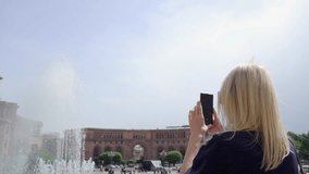 Woman takes photos and videos on her phone against backdrop of Republic Square in Yerevan. Singing fountains and people strolling on hot sunny day. City center, attractions in Yerevan.