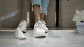 Girl with close-up of her legs and tries on shoes in fashion boutique. She chooses white sneakers, around lot of boxes and shelves with styles. Buying shoes and trying them on.