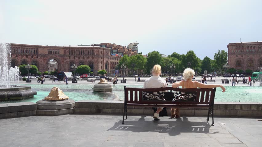 Two women sitting on bench and enjoying pleasant sunny day in city center on Republic Square in Yerevan, Armenia with fountains in backdrop. Enjoying life in beautiful adulthood.