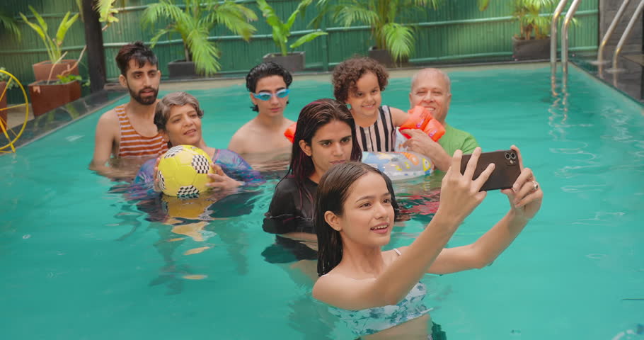 Happy Indian family having fun enjoying outdoor picnic teen girl taking selfie with old senior parents using mobile phone at blue water park. Cute child relax in colorful toy floating ring at resort. Royalty-Free Stock Footage #1106512137