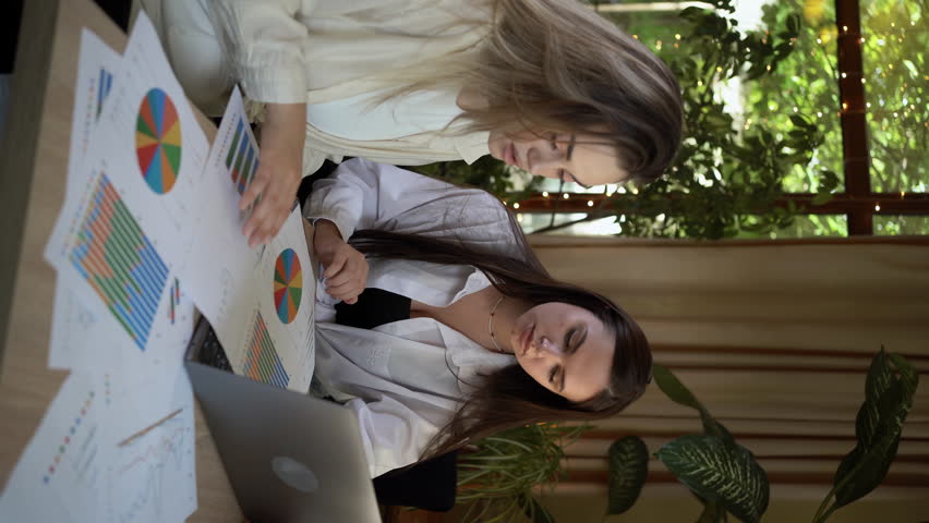 Teamwork, two young businesswomen sitting at table. Women doing financial analysis with a laptop and paper charts at the window of a cafe - Powered by Shutterstock - Get 15% off with code: PIKWIZARD15