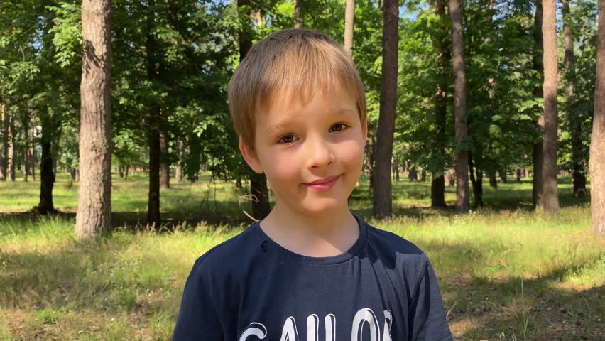 Portrait adorable elementary age boy in the great outdoors on a sunny day. Sweet male child smiling, puts on hat, looking at camera while family camping in the woods. Charming kid poses for best shot