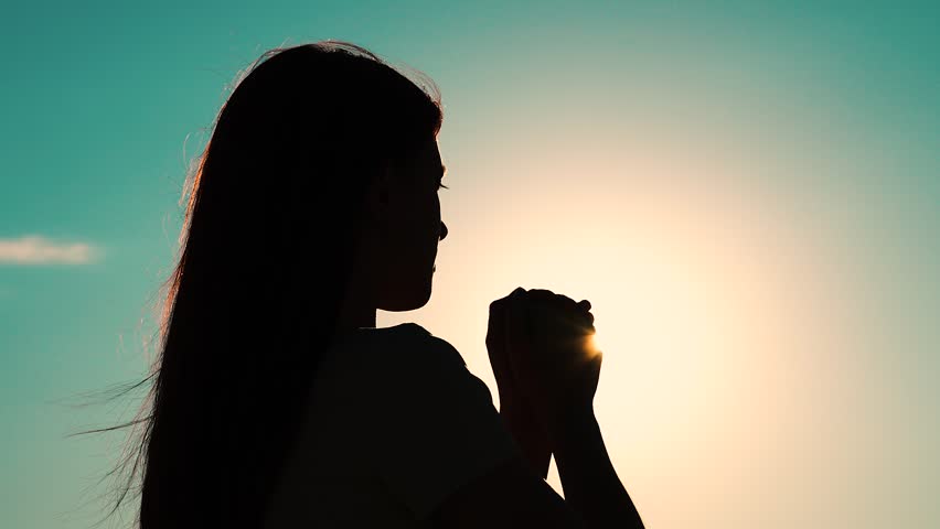 Faith in god. Christian woman praying at sunset close-up. Girl on background of sky in rays of sun prayer to family and children. Relaxation and meditation in nature. Apologize. Hope. Silhouette