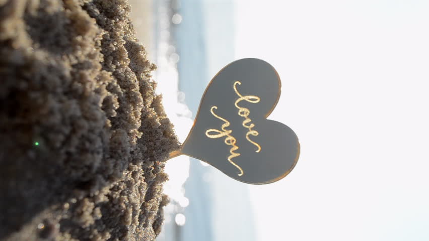Plastic stick shape of heart with words Love you in sand beach of sea shore with sea waves on sunny summer day close-up. Concept love relationship romance amour celebration St. Valentines Day Vertical Royalty-Free Stock Footage #1106532755