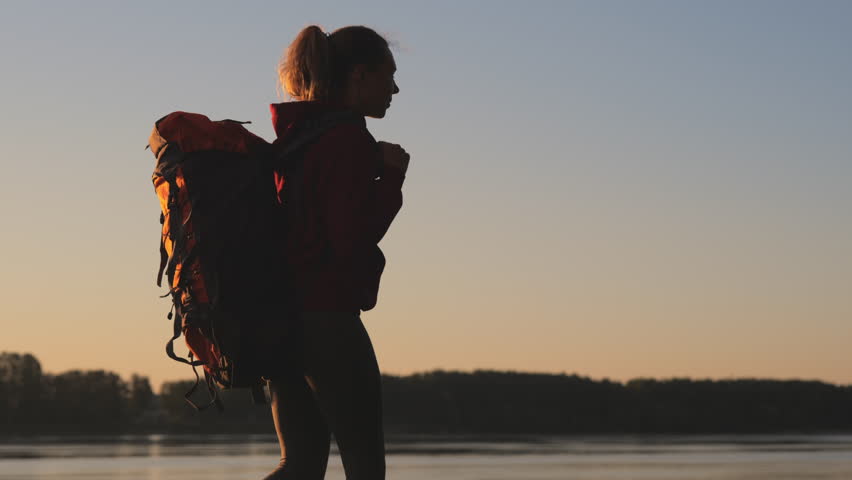 Silhouette of a young woman trekking with a huge backpack along the river at sunset or sunrise. Victory over oneself. Girl enjoys the journey. Concept of hiking, healthy lifestyle, travelling.