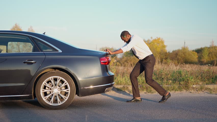 African American man in suit is pushing car along the road. Businessman driver car breakdown. Manager have force majeure, car broke down. Roadside assistance concept. Outdoors. Royalty-Free Stock Footage #1106537339