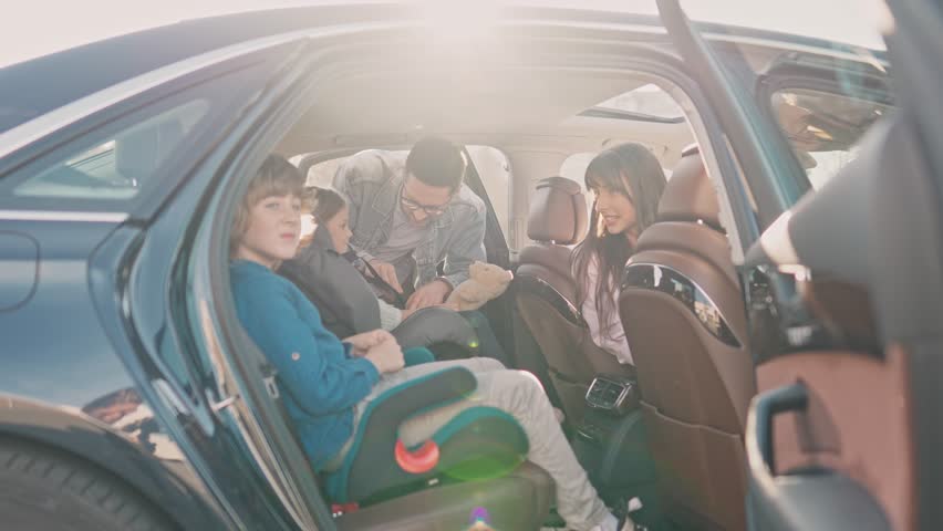 Happy young family is preparing for car trip. Handsome adult man fastens seat belts to two small children. Mother shows concern and encourages children. Caring for safety while traveling on the road. Royalty-Free Stock Footage #1106537377