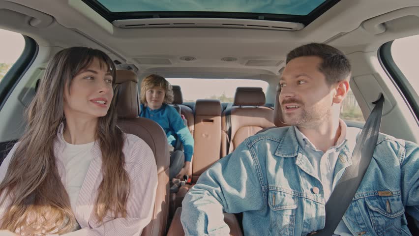 Front view of joyful beautiful parents and their child smiling driving car in the city. Young family having fun together in car in summer. Concept of lifestyle, happy family, travel together.