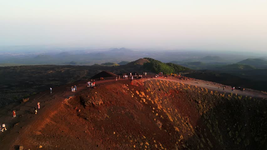 Aerial landscape view of tourists walking on an ancient inactive crater of volcanic magma rock on the Etna volcano mount in Sicily with a view of the city of Catania at sunset lights. Royalty-Free Stock Footage #1106546533
