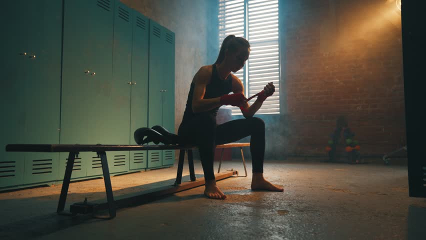 Serious woman fighter boxer wraps hands wrists with boxing bandage sits on bench in locker room, bottom view. Girl kickboxer prepares for fight. Professional sport, competition, preparation concept. Royalty-Free Stock Footage #1106549689
