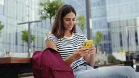 Happy hispanic student girl using yellow cell phone outdoors at University campus. Smiling young woman having fun with online app 