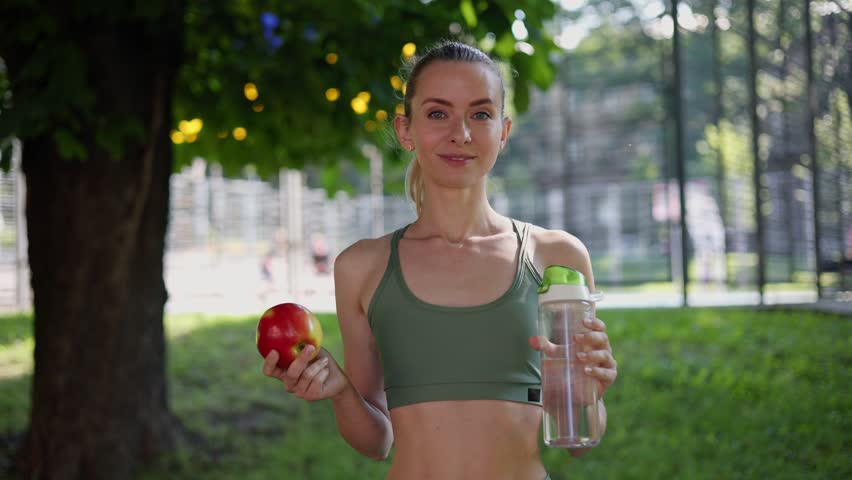 Sporty athletic toned slim girl in sportswear holds a red apple and fresh water in a bottle pointing to a healthy lifestyle. Female healthy athlete looks at the camera with a smile outdoor.