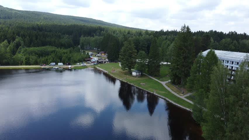 Sunbathing at grassy beach at Milovsky Pond in Protected landscape area Zdarske vrchy in Vysocina region of Czech republic drone panning with resort hotel nearby