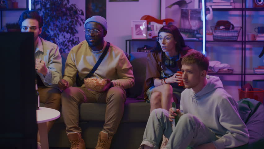 Full shot of young woman and three diverse male friends sitting on couch at home on Friday night, drinking beer with crisps, anxiously watching sport match on TV and shaking head in disappointment Royalty-Free Stock Footage #1106582329